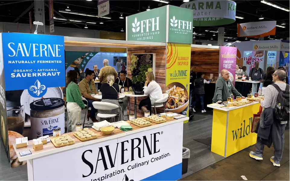 20x30 custom modular island booth at NPE West featuring Saverne, Wildbrine, and FFH branding with sampling counters and seated meeting space.